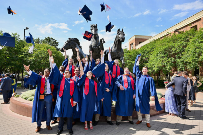 A group of class of 2023 graduates toss their caps in the air in front of the Moody Coliseum Mustang statues. Image courtesy SMU.