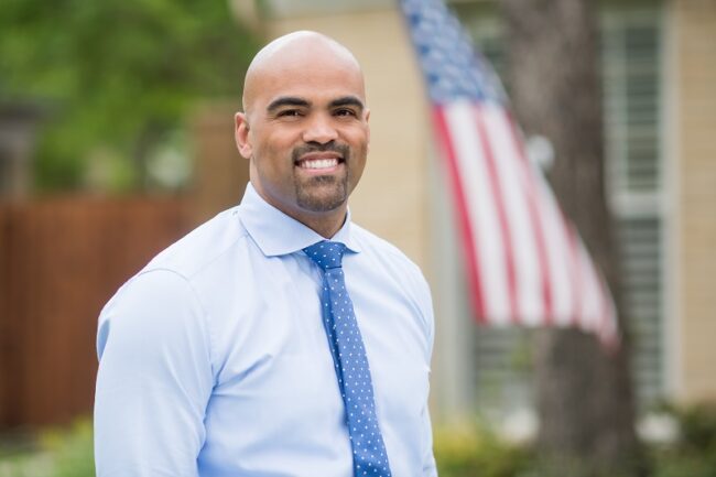Colin Allred stands in front of a building with an American flag out front. Photo credit: Colin Allred for Congress