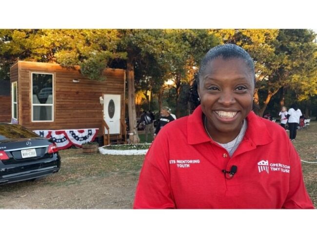 A smiling woman in a red polo outdoors in front of a wooden tiny house.