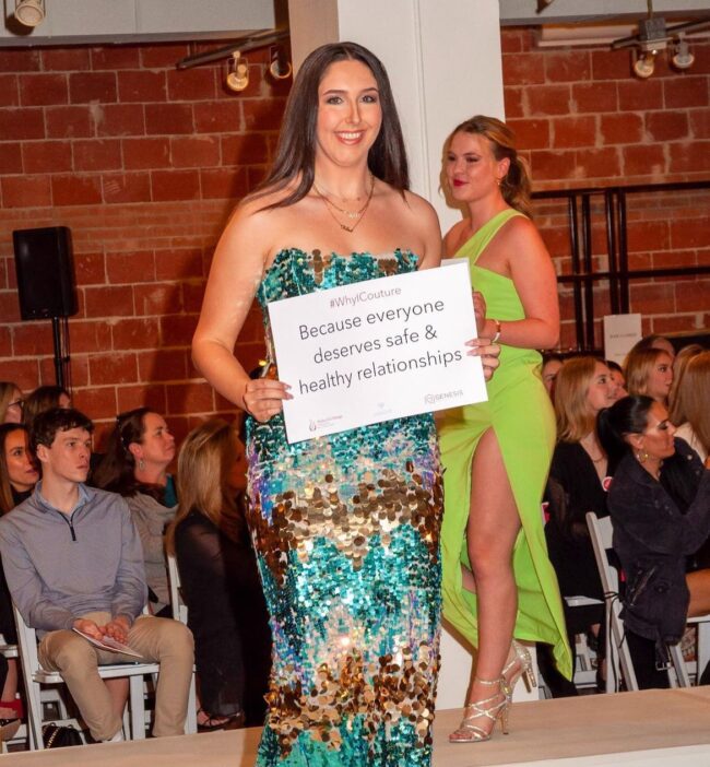 A model sporting fashions from Tootsies walks the runway at Alpha Chi Couture holding a sign representing why she chose to walk in the show to support domestic violence survivors. Image courtesy of SMU Alpha Chi Omegas Instagram.
